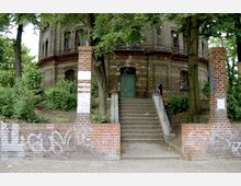 Backsteinbau mit Rundbogenfenstern und einer grünen Tür, die über eine Treppe mit seitlichen Geländern erreichbar ist. Im Vordergrund steht eine Ziegelmauer mit zwei Säulen, umgeben von Bäumen und dichtem Grün.