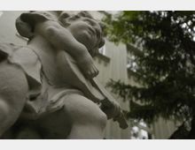 Eine Nahaufnahme einer steinernen Engelsskulptur, die eine Harfe hält, mit einem unscharfen Hintergrund aus einem hellen Gebäude und Nadelbaumästen.