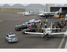 Flughafenszene mit mehreren geparkten Kleinflugzeugen vor Flugzeughangars. Im Vordergrund befinden sich Polizeifahrzeuge, ein Sportwagen und mehrere Personen, die an einer scheinbar filmischen Inszenierung mit Kameratechnik und Equipment arbeiten.