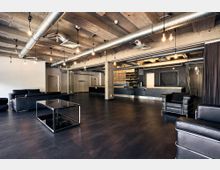 Industrie-inspirierter Innenraum mit freiliegenden Betonwänden und sichtbaren Lüftungsrohren an der Decke. Der Raum ist mit schwarzen Ledermöbeln, einem dunklen Holzboden und einer Theke aus Holz und Metall gestaltet, ergänzt durch minimalistische Hängelampen.