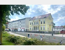 Straßenszene mit einem modernen grauen Wohn- und Geschäftsgebäude sowie einer gelben Altbauvilla mit dekorativen Fassadenelementen und Balkon. Im Vordergrund verläuft ein Fußweg entlang einer betonierten Mauer mit Graffiti, umgeben von Büschen und Bäumen.