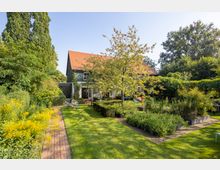 Ein Garten mit gepflegtem Rasen, Zierpflanzen und einem gepflasterten Weg, der zu einem Haus mit dunkler Holzfassade und rotem Ziegeldach führt. Im Vordergrund stehen ein Baum, Büsche und Blumenbeete, umgeben von hoher grüner Vegetation.