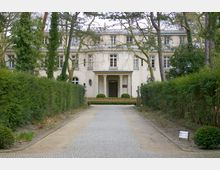 Helle, historische Villa mit drei Stockwerken, säulengestütztem Eingang und symmetrischen Fenstern, umgeben von gepflegten Hecken, Bäumen und einem Weg aus Kopfsteinpflaster, der auf das Gebäude zuführt. Im Vordergrund stehen zwei steinerne Statuen an den Seiten des Weges.