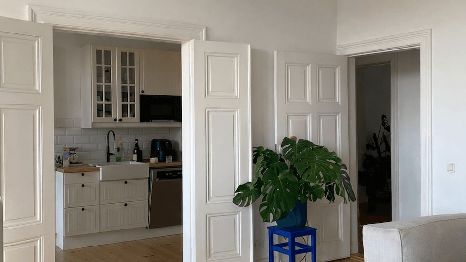 Bright, minimalist living room with white paneled double doors opening into a small white kitchen with tile backsplash and light wood floors. A large circular ceiling light, a gray area rug, and a potted plant on a blue stool sit near a doorway leading to another room.