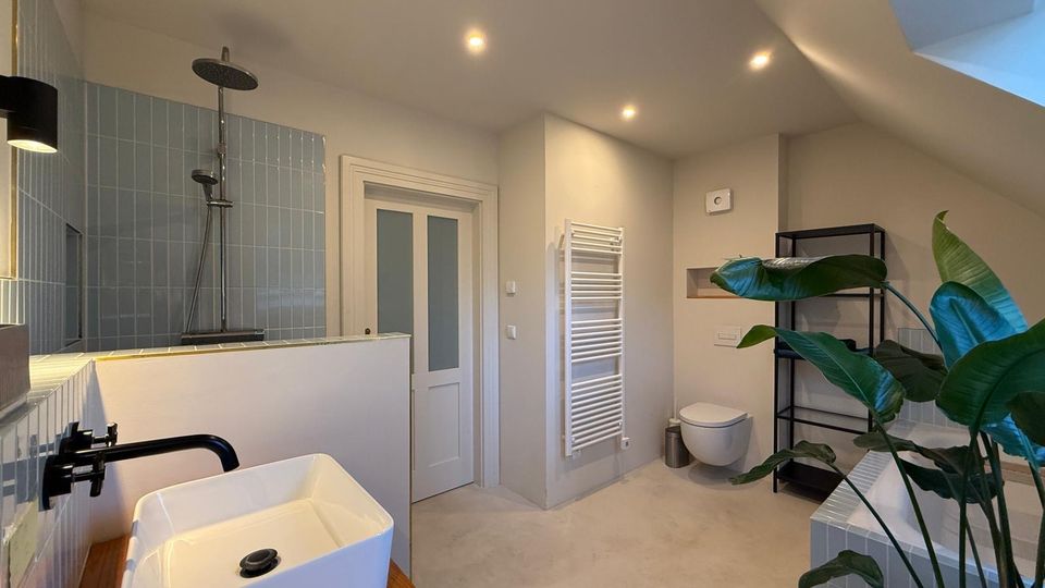 Modernes Badezimmer mit einer bodengleichen Dusche, hellblauen Wandfliesen, einem freistehenden Waschbecken auf einer Holzplatte und einem weißen WC. Der Raum ist mit einem Heizkörper, einer Pflanzen-Dekoration und einem schwarzen Regal ausgestattet.