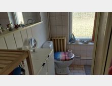 Kleines, gefliestes Badezimmer mit WC, weißem Unterschrank und Wandregal mit Spiegel. Daneben ein großes Fenster mit Milchglas, darunter eine geflieste Ablagefläche sowie ein Heizkörper in der Ecke.