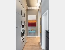 Schmaler, moderner Küchenflur mit hellen Einbauschränken und integrierten Backöfen links, Holzfußboden und grauen Wänden. Am Ende hängt ein abstraktes Bild über einem gläsernen Konsolentisch; die Decke zeigt dekorativen Stuck und eine lineare Deckenleuchte.