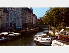 Flusskanal mit anlegenden Booten und historischen Wohngebäuden am Ufer; rechts ein Baumsaum und eine Promenade mit Sitzbereichen, im Hintergrund eine kleine Brücke.