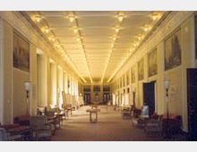 Langer, schmaler Innenraum mit hoher Decke und symmetrischem Design, beleuchtet durch eine Reihe von Deckenleuchten. Die Wände sind mit Gemälden dekoriert, und entlang des Raumes stehen Stühle und Tische in regelmäßigen Abständen.