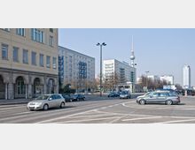 Straßenszene in einer Stadt mit mehrspuriger Fahrbahn und mehreren Autos, umgeben von modernen Wohngebäuden. Im Hintergrund ist der Fernsehturm zu sehen, ein bekanntes Wahrzeichen, sowie weitere Hochhäuser.