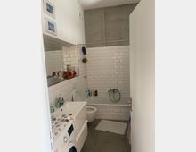 Badezimmer mit weiß gefliesten Wänden und einer Badewanne. Im Vordergrund befinden sich ein Waschbecken mit Spiegel sowie eine Waschmaschine, während im Hintergrund die Toilette und Duschutensilien zu sehen sind.