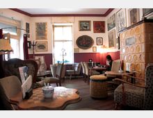 Gemütliches Café mit retro-inspiriertem Interieur, das aus Polstersesseln, Holztischen und einer gelben Kachelheizung besteht. Die Wände sind mit Gemälden und Kunstwerken dekoriert, während durch ein Fenster Tageslicht einfällt.