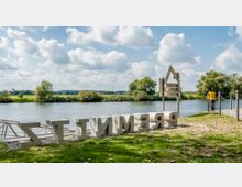 Flussufer mit einer Wiese im Vordergrund, auf der ein Schriftzug aus großen, liegenden Buchstaben steht. Im Hintergrund ist ein ruhiger Fluss mit Bäumen und Wiesen zu sehen, darüber ein blauer Himmel mit lockeren Wolken.