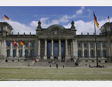 Frontansicht des Reichstagsgebäudes in Berlin mit seiner markanten Glaskuppel, Säulenfassade und der Inschrift „Dem Deutschen Volke“. Im Vordergrund befinden sich mehrere Flaggen, Stufen führen zum Eingangsbereich, und Menschen sind auf dem Platz und den Treppen verteilt.