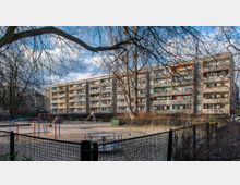 Ein mehrstöckiges Wohngebäude im Plattenbaustil mit Balkonen, das von Bäumen umgeben ist. Im Vordergrund befindet sich ein umzäunter Spielplatz mit Spielgeräten wie einer Wippe, einer Rutsche und einem Karussell.