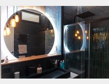 Modernes Badezimmer mit dunklen Wandfliesen und einer großen, runden Spiegelbeleuchtung. Zu den sichtbaren Elementen gehören ein schwarzes Waschbecken mit Armatur, eine Glasdusche mit Regenduschkopf, hängende Glühbirnen als Beleuchtung sowie ein schmaler Holzablagebereich unter dem Spiegel.