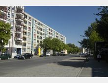 Eine breite Straße in einer urbanen Umgebung mit mehrstöckigen Wohngebäuden auf der linken Seite, großen Fenstern und Balkonen. Neben der Straße sind parkende Autos, Bäume und ein Bürgersteig sichtbar; im Hintergrund weitere Gebäude und blauer Himmel.