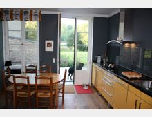 Moderne Küche mit gelben Unterschränken, schwarzer Arbeitsplatte und Dunstabzugshaube. Im Vordergrund steht ein runder Holztisch mit Stühlen, und durch die große Glastür im Hintergrund ist ein Garten sichtbar, in dem ein schwarzer Hund steht.