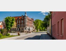 Straßenszene in einer Kleinstadt mit einem historischen Gebäude aus rotem Backstein und einem Turm im Vordergrund, flankiert von weiteren Gebäuden mit farbigen Fassaden. Im Hintergrund sind grüne Bäume und ein blauer Himmel mit wenigen Wolken sichtbar.