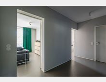 Moderner, schlichter Innenraum mit grauem Boden und Wänden, der einen Flur mit offenen Türrahmen zeigt; durch eine Tür sieht man ein Zimmer mit schwarzem Ledersofa, grünen Vorhängen und einem weißen Schrank.
