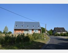 Modernes Wohnhaus mit Holzfassade und steilem Dach aus Wellblech, das vier rechteckige Fenster aufweist. Das Gebäude steht in einer ländlichen Umgebung mit Büschen und einer Nebenstraße im Vordergrund.