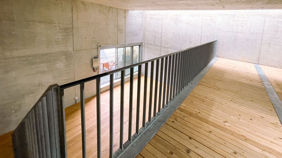 Ein minimalistischer Innenraum mit Betonwänden und einer Decke aus Sichtbeton, einer Holzplattform mit Metallgeländer, sowie einer bodentiefen Glasfront, die Tageslicht hereinlässt.