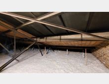 Blick in ein Dachgeschoss mit Metallträgern, einer Ziegelmauer und einer gedämmten Fläche aus weißem Material. Im Vordergrund sind dünne Holzstäbe und einzelne senkrecht stehende Markierungen zu sehen.