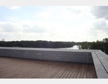 Eine Dachterrasse mit Holzboden und niedriger grauer Steinmauer, die den Blick auf eine grüne Baumlandschaft und einen Fluss freigibt. Im Hintergrund ist ein Riesenrad und der bewölkte Himmel zu sehen.