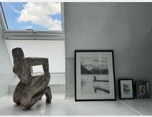 Ein minimalistischer Innenraum mit schräger weißer Fliesenwand, einem Dachfenster mit halb geschlossener Jalousie und blauem Himmel im Hintergrund. Im Vordergrund stehen eine abstrakte Holzskulptur in menschlicher Form und gerahmte Schwarz-Weiß-Fotografien, die verschiedene Motive zeigen.