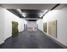 Ein moderner Ausstellungsraum mit weißen Wänden und schwarzem Boden, an den Wänden hängen verschiedene Kunstwerke. In der Mitte des Raumes stehen weiße Stühle, und die Decke ist mit Neonröhren beleuchtet.