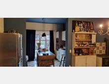 Küche mit einer zentralen Kücheninsel aus Holz, großen Fenstern mit dunkelblauen Vorhängen im Hintergrund und einem Fliesenboden mit geometrischem Muster. Rechts befindet sich ein heller Schrank mit Gläsern und Flaschen darauf sowie eine Wand mit dekorativen Fliesen neben einem Kamin.