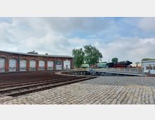 Rundlokschuppen mit roten Backsteinfassaden und geschlossenen Toren, davor Gleise und ein Drehteller. Rechts im Bild steht eine schwarze Dampflok sowie ein grüner Waggon, umgeben von Bäumen und freiem Himmel.