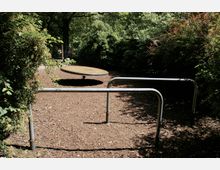 Spielplatz mit einer runden Drehscheibe aus Metall auf einer Fläche aus Rindenmulch, umgeben von hohen Büschen und Bäumen. Im Vordergrund sind Metallgeländer, die den Bereich abgrenzen.