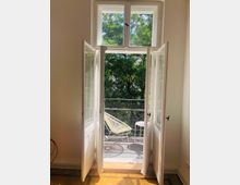 Weiß gestrichene Balkontüren öffnen sich zu einem kleinen Balkon mit einem Metallgeländer, darauf stehen ein heller, filigraner Stuhl und ein kleiner Tisch. Im Hintergrund sind dichte grüne Bäume sichtbar, während Sonnenlicht durch das Fenster auf den Holzboden des Innenraums fällt.