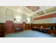 Historischer Büroraum mit Holzvertäfelung, bogenförmigen Türen und dekorativem Stuck an der Decke. Ein langer Holztisch mit Stühlen und Arbeitsplätzen sowie eine Sitzbank dominieren den Raum, der durch Wandlampen und Pendelleuchten beleuchtet wird.