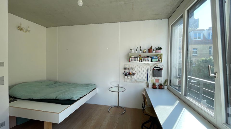 Minimalistisch eingerichtetes Zimmer mit Beton- und Holzelementen, einem schwebenden Bett links, einem langen weißen Schreibtisch vor einem großen Fenster rechts und Regale sowie Wandpaneele mit kleinen Pflanzen und Schreibutensilien an der Wand.
