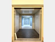 Blick aus einem offenen Lastenaufzug in einen Flur mit hellgrauen Wänden und Türen. Am Ende des Flurs befinden sich zwei geschlossene Türen, die mit den Beschriftungen „Lager Shop: 1“ und „Aufsichtsraum“ versehen sind.