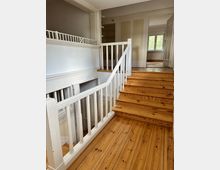 Holztreppe in einem hellen Wohnraum mit weißem Geländer und Dielenboden aus Holz. Im Hintergrund sind Türen, Wände in neutralen Farben sowie ein Fenster mit Blick ins Freie zu sehen.