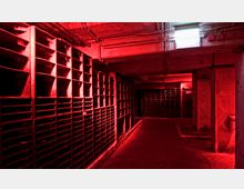 Industrieartiger Raum mit offenen Metallregalen an den Wänden, beleuchtet durch rotes Licht. Sichtbare Rohrleitungen verlaufen an der Decke, und vereinzelt sind Graffiti an den Wänden erkennbar.