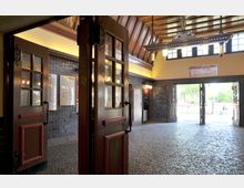 Eingangsbereich eines historischen Gebäudes mit offenen Holztüren und Glasfenstern, blau gefliesten Wänden und einem mit Kopfsteinpflaster belegten Boden. Im Hintergrund führen weitere Türen nach draußen, wo Bäume und ein sonniger Straßenbereich sichtbar sind.