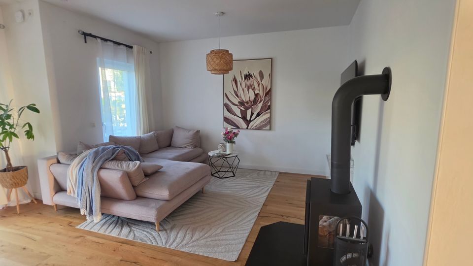Helles, modern eingerichtetes Wohnzimmer mit L-förmigem Stoffsofa auf hellem Teppich, kleinem Beistelltisch und großem Blumenbild an der Wand. Links sorgt ein Fenster mit weißen Vorhängen für Tageslicht, rechts steht ein schwarzer Kaminofen mit Ofenrohr auf Holzboden.