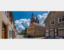 Straßenszene in einer historischen Altstadt mit gepflastertem Weg, flankiert von mehrstöckigen Häusern mit verputzten Fassaden und roten Ziegeldächern. Im Hintergrund ragt eine Kirche mit hohem Turm und spitzem Dach vor einem Himmel mit weißen Wolken hervor.