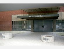 Moderne Kirchenfassade aus rotem Backstein und Beton der evangelischen Christuskirche mit breitem Vordach; im unteren Bereich Glasbausteine und dunkelgraue Eingangstüren. Im Vordergrund stehen runde Betonpflanzkübel mit Graffiti.