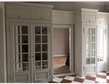 Weiß gestrichene Einbauschränke mit Glastüren und integrierten Spiegeln flankieren eine offene Tür, die zu einem Raum mit Parkettboden führt. Der Boden im Vordergrund besteht aus diagonalen Fliesen in Weiß und Rotbraun.