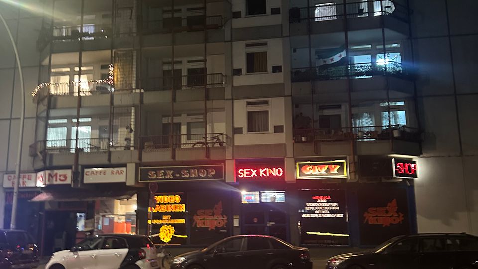 Mehrstöckiges Wohn- und Geschäftsgebäude bei Nacht, mit beleuchteten Balkonen in den oberen Etagen und Gewerbeeinheiten im Erdgeschoss, darunter ein Café, ein Sexshop und ein Sexkino mit leuchtenden Neon-Schildern. Vor dem Gebäude parken mehrere Autos entlang einer zweispurigen Straße.