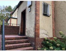 Gelbes Gebäude mit bröckelndem Putz, rotem Backsteinsockel und einem kleinen Fenster mit Metallgitter. Eine rote Backsteintreppe mit Geländer führt zu einer Eingangstür, daneben sind grüne Büsche zu sehen.