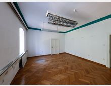 Leerer, heller Innenraum mit Fischgrät-Parkettboden, weißen Wänden mit türkisfarbener Zierleiste und großen rechteckigen Deckenleuchten. Links befindet sich ein hohes Fenster über einem Heizkörper, gegenüber eine weiße Tür und rechts ein weiterer Türdurchgang.