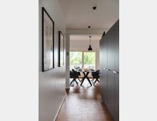 Flur mit Holzfußboden, an dessen Ende ein Essbereich mit einem runden Tisch, vier Stühlen und einer großen Fensterfront mit Blick ins Grüne sichtbar ist. Links hängen gerahmte Bilder an einer weißen Wand, rechts befindet sich eine graue Einbauschrankwand.