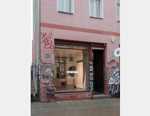 Fassade eines Altbaus mit rosa Anstrich, großen Fenstern und Graffitis auf den unteren Wänden. Im Schaufenster ist ein Fotostudio sichtbar, beleuchtet von einem Leuchtschild mit der Aufschrift "FOTOSTUDIO" und ausgestattet mit professionellem Studio-Equipment.