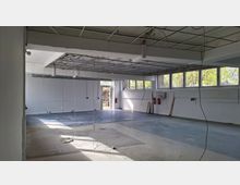 Großer, leerer Innenraum einer ehemaligen Werkstatt oder Lagerhalle mit Betonboden, weißen Wänden und abgehängter Decke im Umbau, aus der Kabel herabhängen. Entlang der rechten Seite befindet sich eine Reihe hoher Fenster mit Blick auf Bäume; hinten ist eine offene Tür nach außen sichtbar.